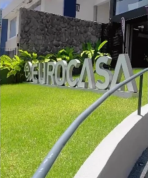 Letreiro “EUROCASA” sobre gramado e jardim tropical, na entrada do evento.