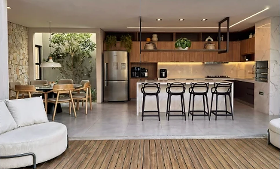 Vista ampla da área gourmet integrada à cozinha com ilha: parede de pedra, jardim interno, marcenaria amadeirada e deck de madeira.